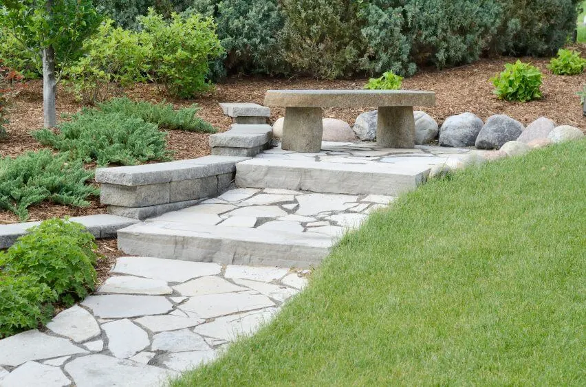 The solitary concrete stone bench atop the concrete stoned pathway is immediately surrounded by rounded stones on one side and curved paving slabs on the other The patches of green shrubs and the sprawling Bermuda grass all seem to be in harmony with the presence of the concrete This garden invites for meditation or simply just some quiet reflections