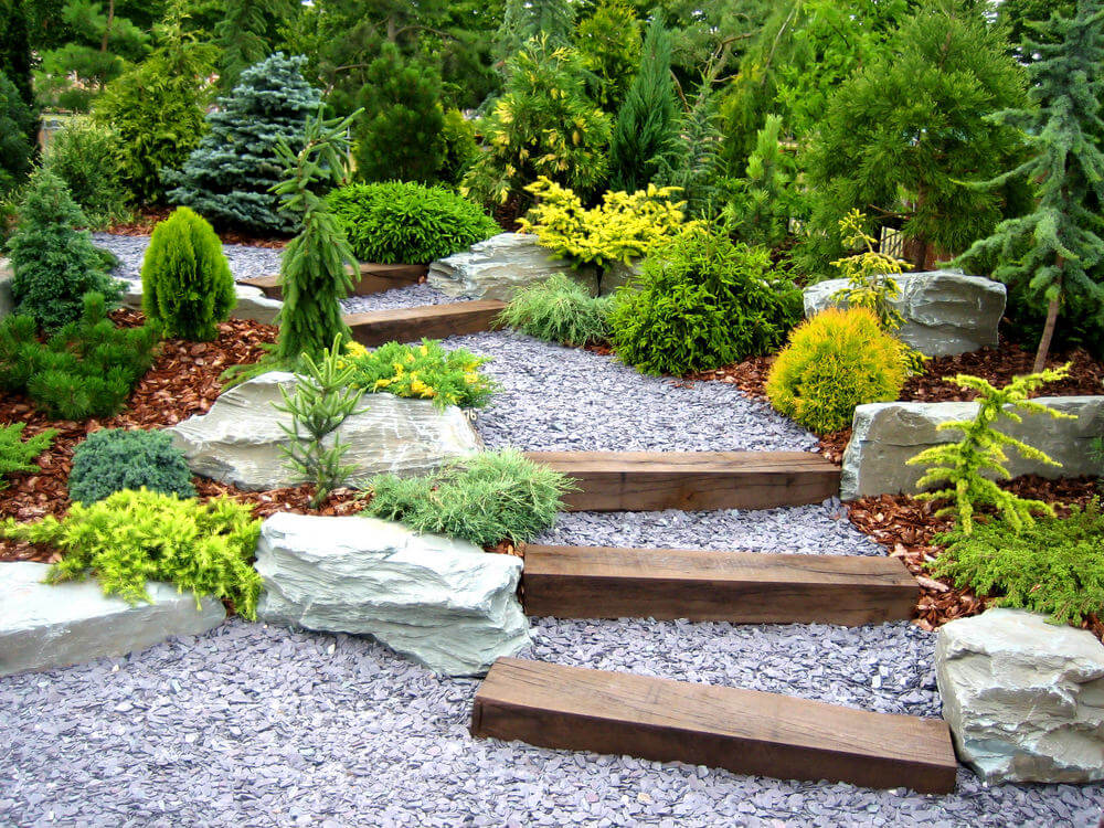 A well-trimmed variety of evergreen shrubs makes way for the garden steps of pebbled stones and occasional wooden slabs Some bleach colored boulders are also used in between the shrubs to allow for enough space