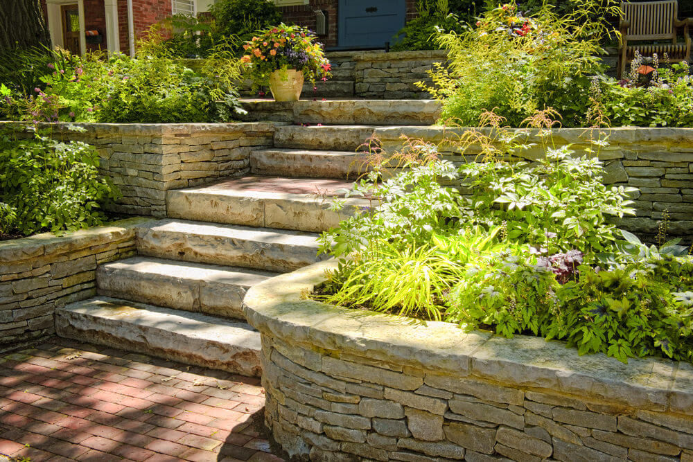 Sunshine greets the garden shrubs that are harmoniously tucked in these landscaped stone-styled planters juxtaposed with concrete garden steps and brick floor