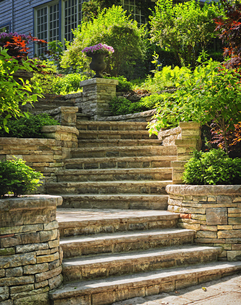 A large archaic vase of pink flowers stands in the center of it all as stone-styled planters tuck away green shrubs to make way for the wide garden steps leading to this bend where the pink flowers are the central attraction
