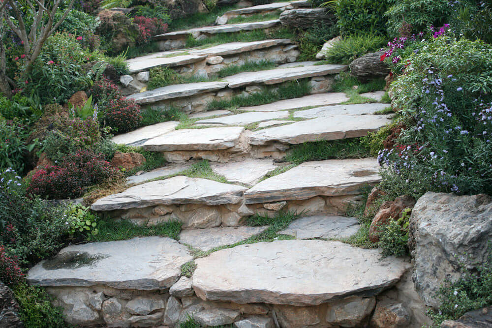 Flowery shrubs seem to usher in guests toward these huge pieces of stone slabs that serve as garden steps where grasses have found their way through the cracks within