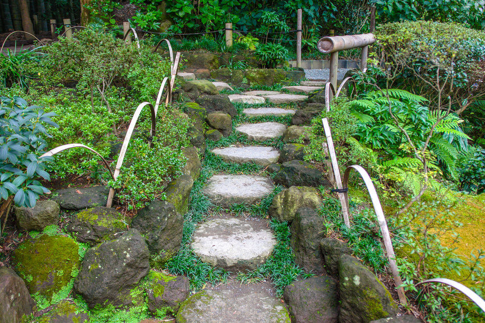 Dark mossy boulders together with bamboo slats and bamboo shoots serve as dividers, separating the ferns and shrubs from garden steps made up of slabs of stone
